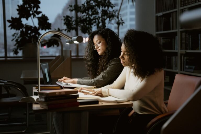 Two women studying in a library with laptops and books.