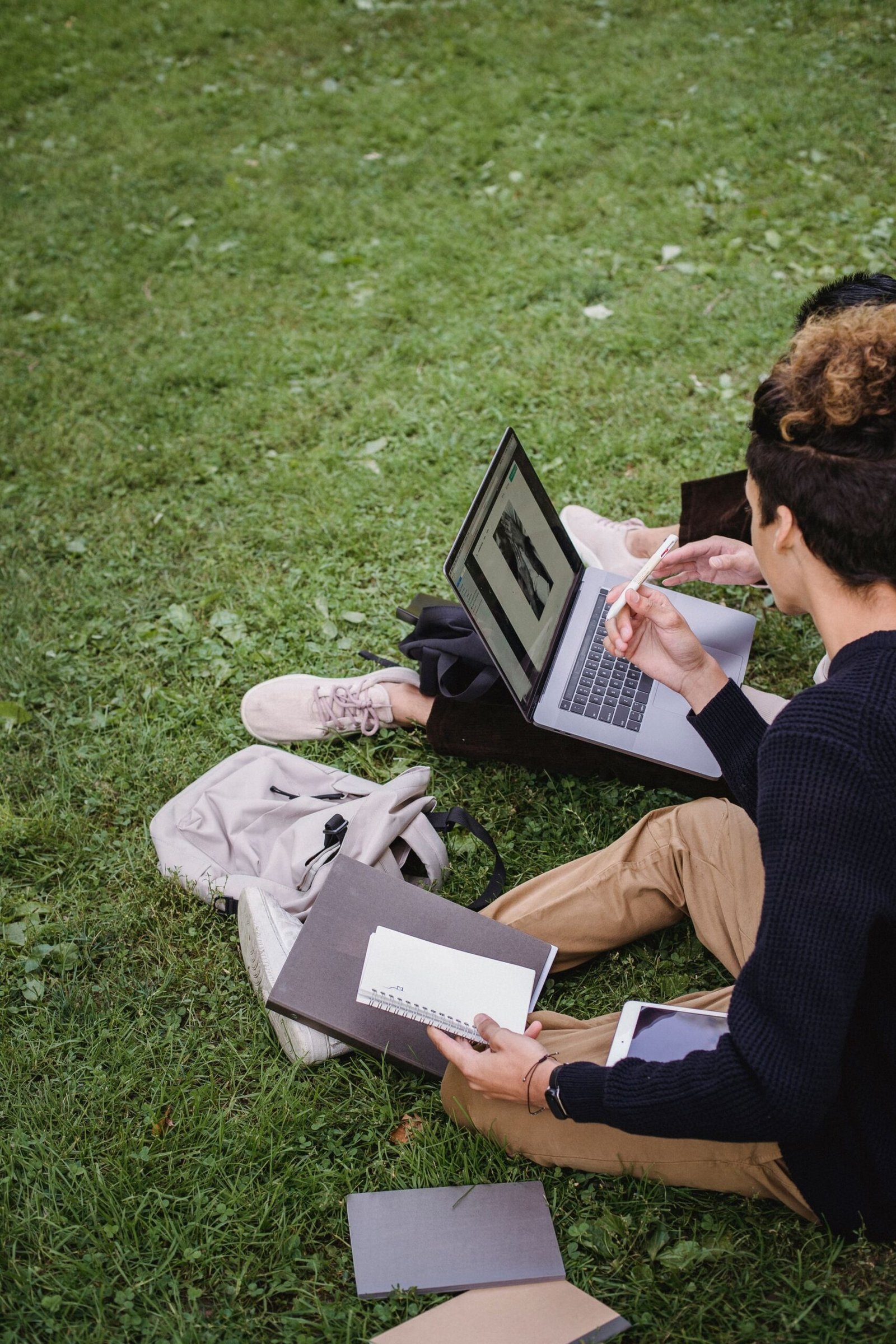 Two students studying together on a laptop outdoors, surrounded by books.