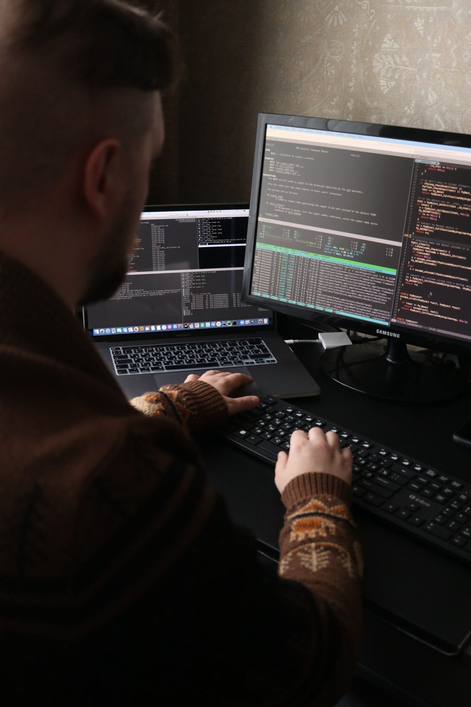 A programmer working on code with dual monitors and a laptop in a dimly lit room.