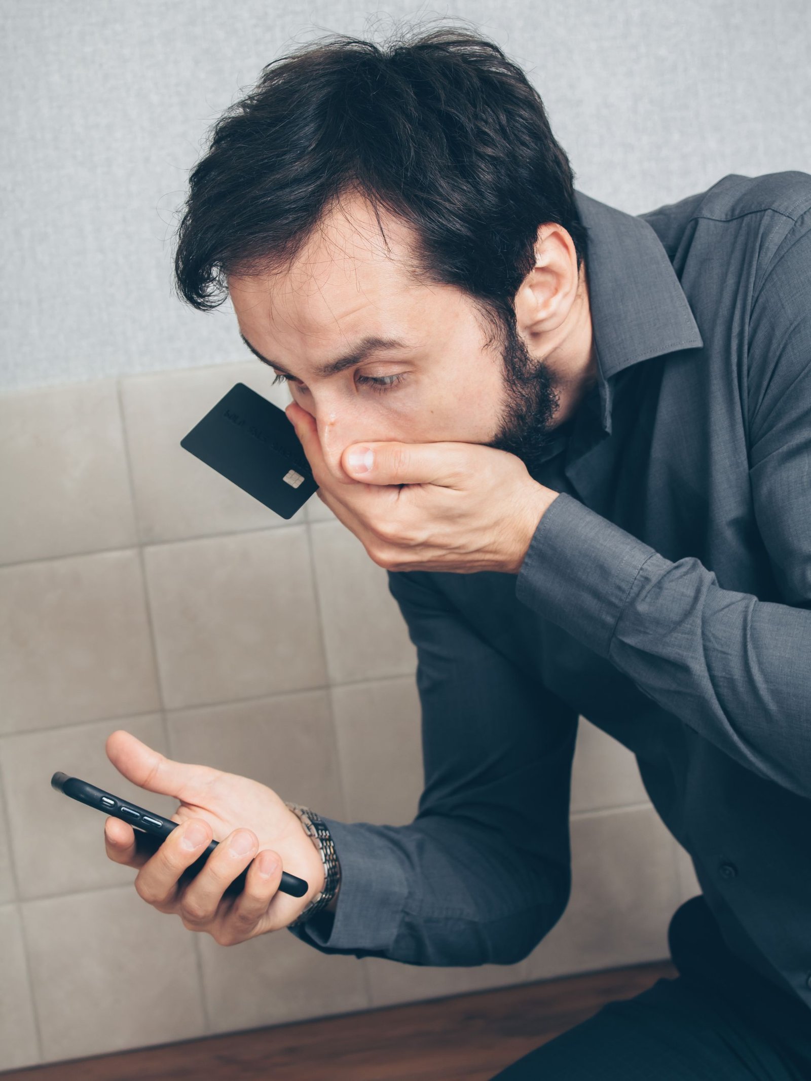Shocked man holding a smartphone and credit card after discovering possible fraud.