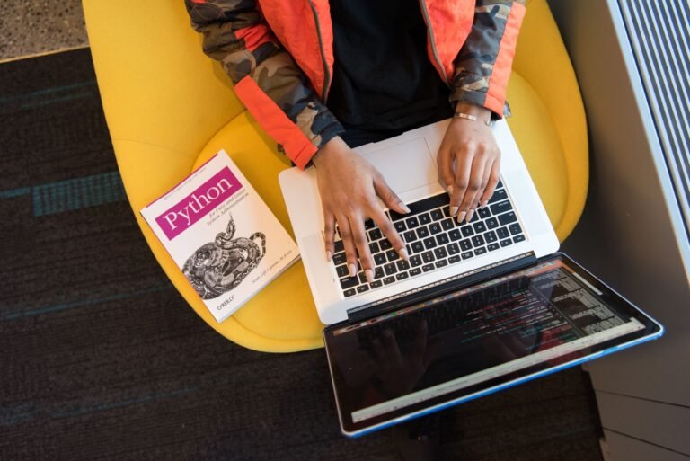 A developer typing Python code on a laptop with a Python book beside her.