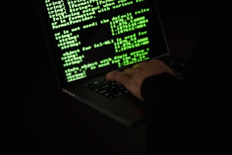 Silhouette of a hooded figure typing on a laptop in the dark with green code glowing on the screen.