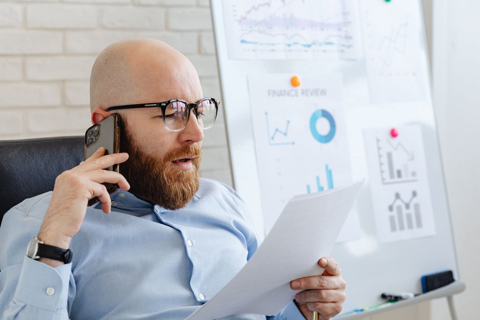 A bearded businessman talking on a smartphone while reviewing documents in an office.