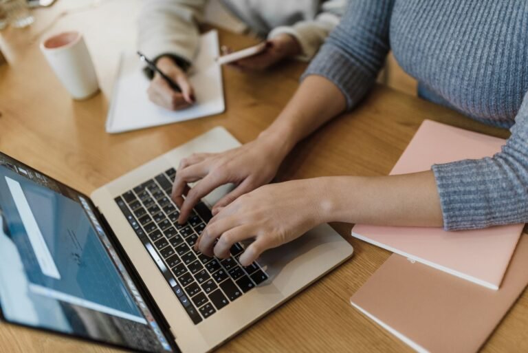 A freelancer typing on a laptop at a wooden desk with coffee and a notebook nearby.