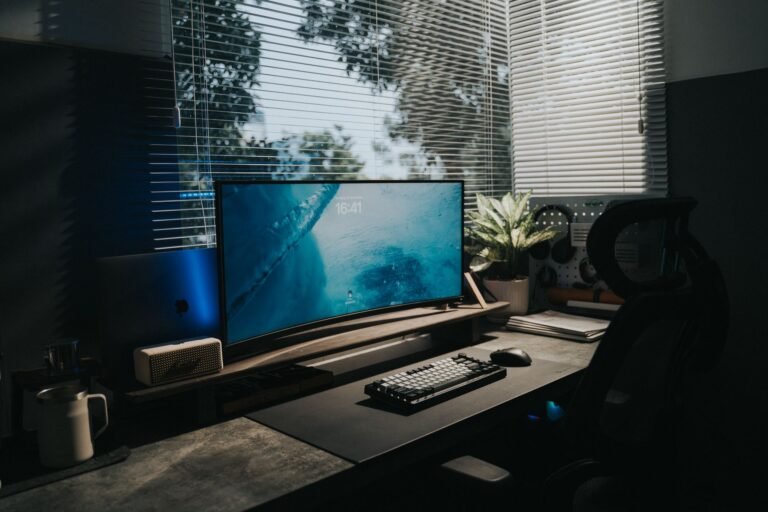 A clean home office desk with a large monitor, keyboard, and a plant by a window.