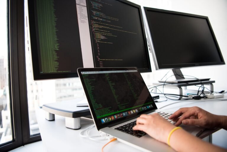 A developer writing code on a laptop in front of multiple monitors in an office setting.